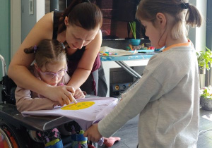 mama pomaga córce na wózku narysować słonko na bluzce a druga córka trzyma i naciąga bluzkę